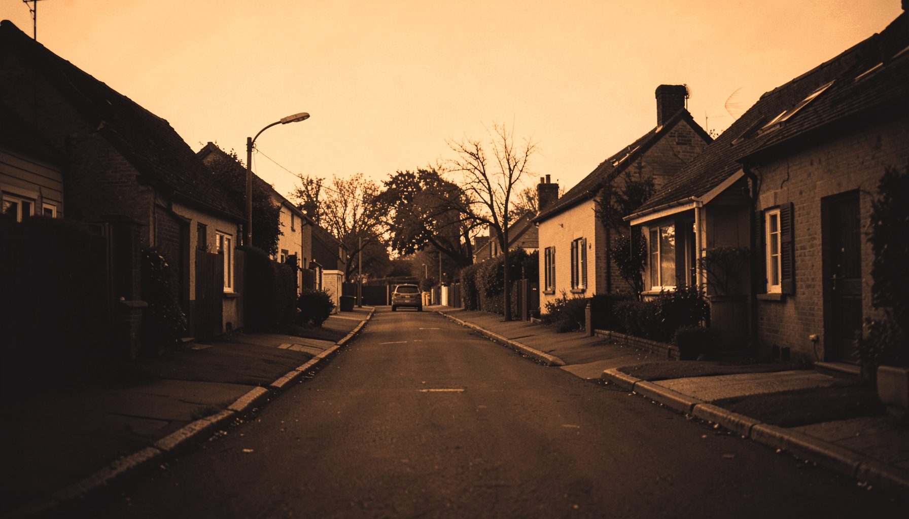 A warm duotone photograph of a quiet residential street with modest houses and soft morning light, rendered in deep amber and charcoal tones.